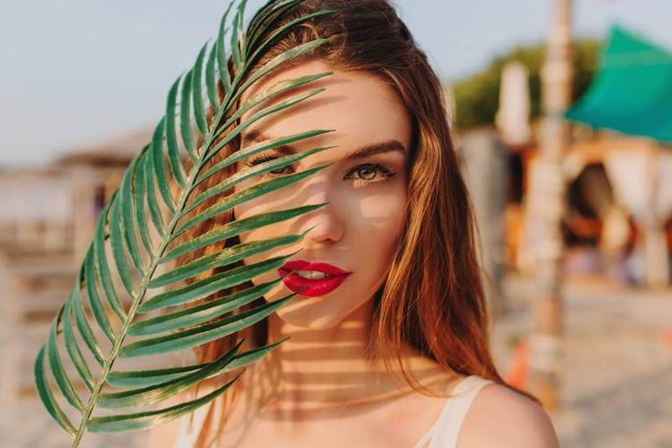 Headshot of a beautiful brunette women in the sun holding a palm leaf over one side of her face
